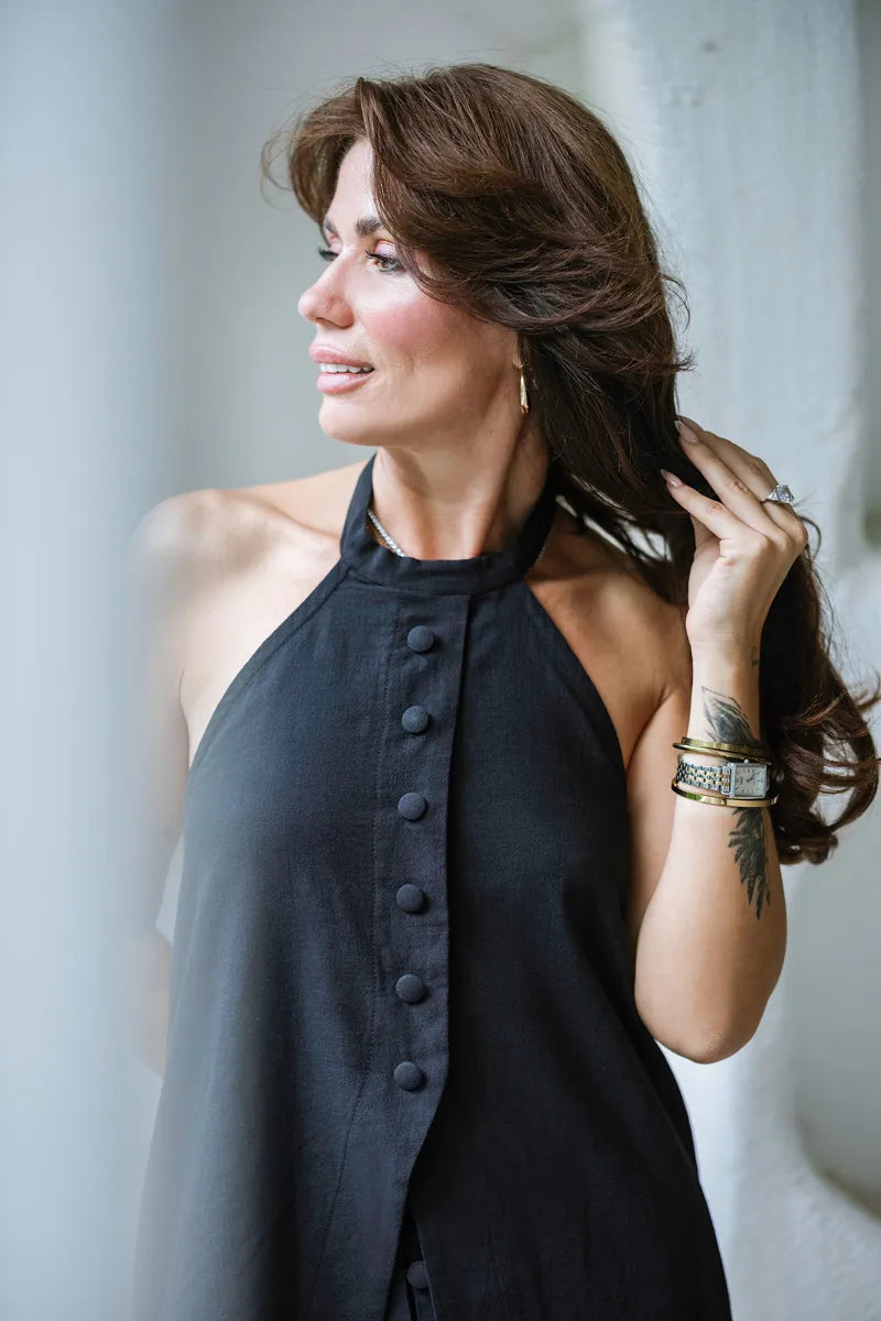 Woman wearing a black sleeveless top with buttons, standing against a light-colored wall.