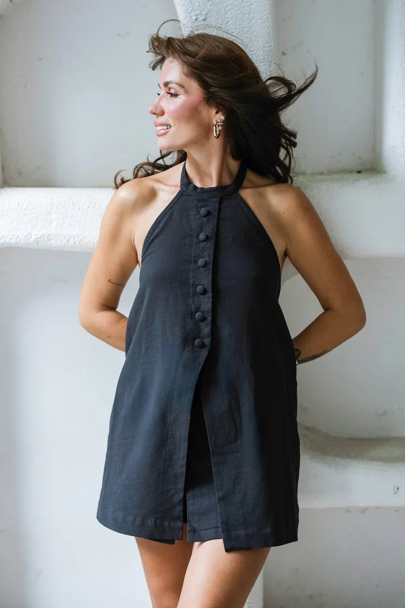 Woman wearing a black sleeveless dress with buttons standing against a white wall.