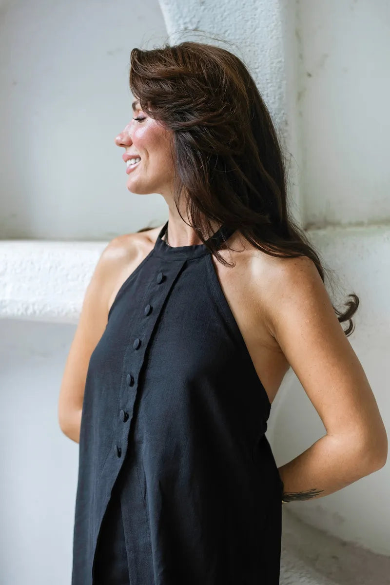 Woman wearing a black sleeveless top with buttons, standing against a white wall.