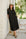 Woman in a black shirt dress and hat standing against a stone wall with greenery in the background