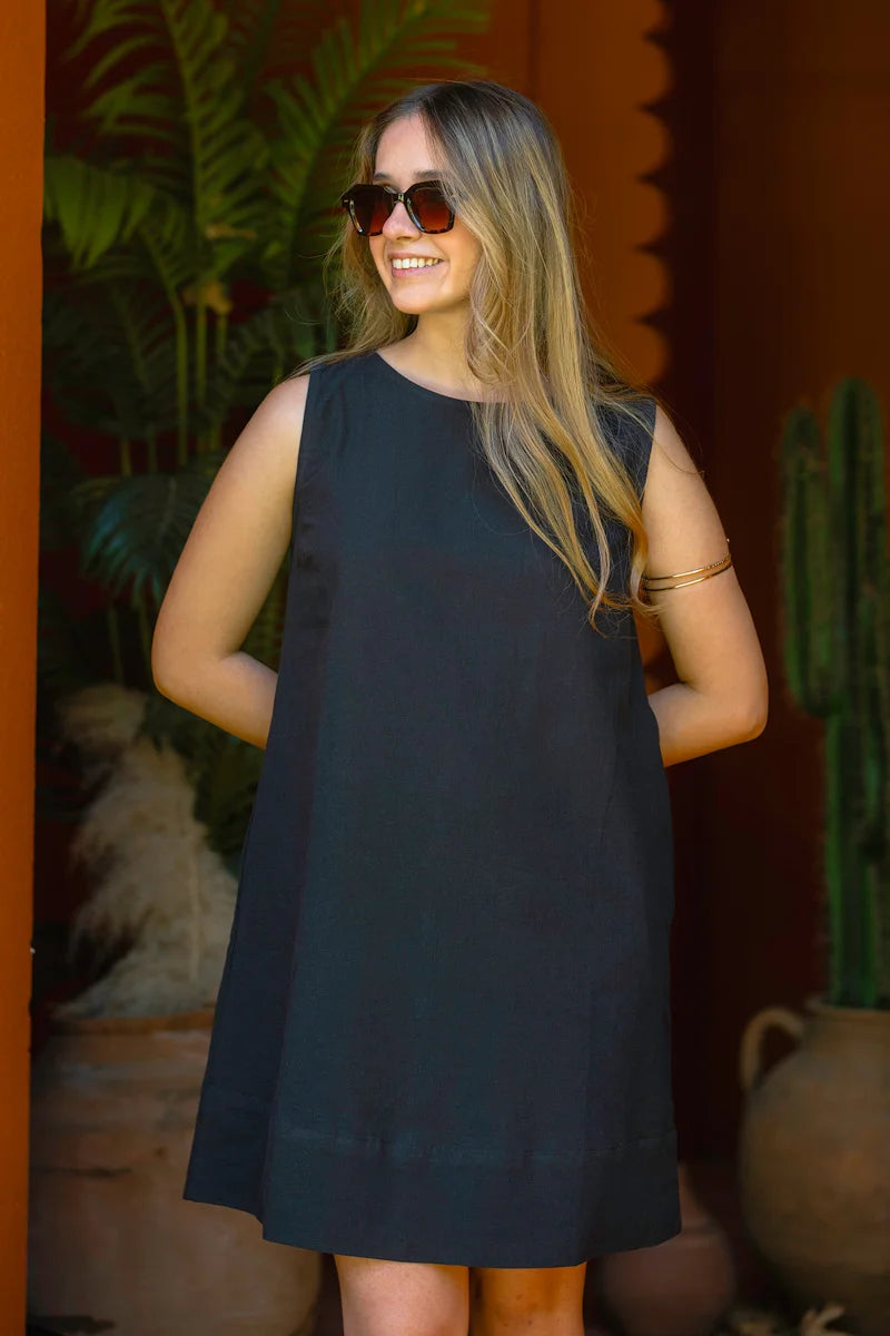 Woman wearing a black sleeveless a line dress with sunglasses, standing against a warm-toned wall with plants.