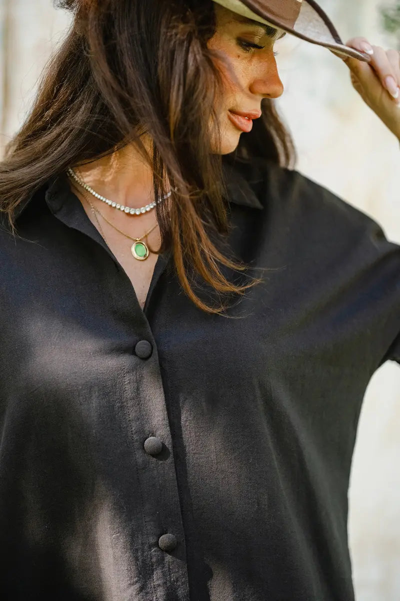 Woman wearing a black shirt dress with buttons, looking down, with a blurred background.