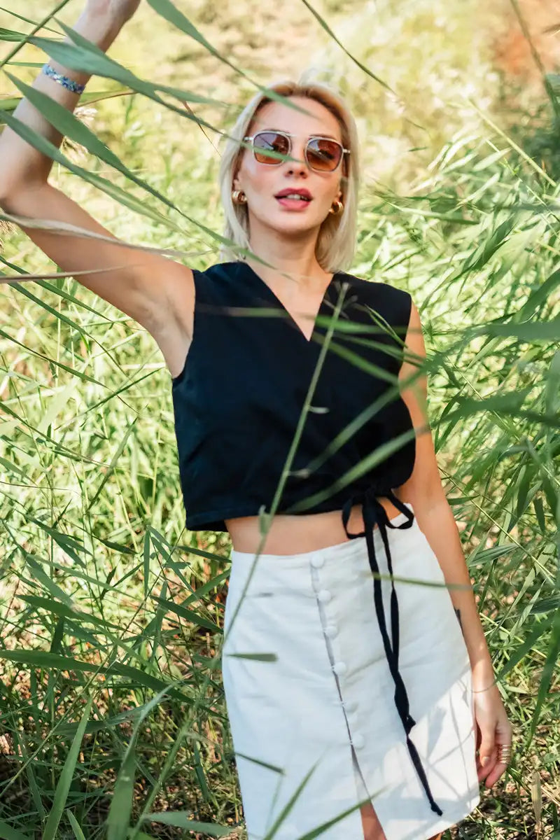 Woman in black sleeveless linen vest top and white skirt standing among tall green grass.