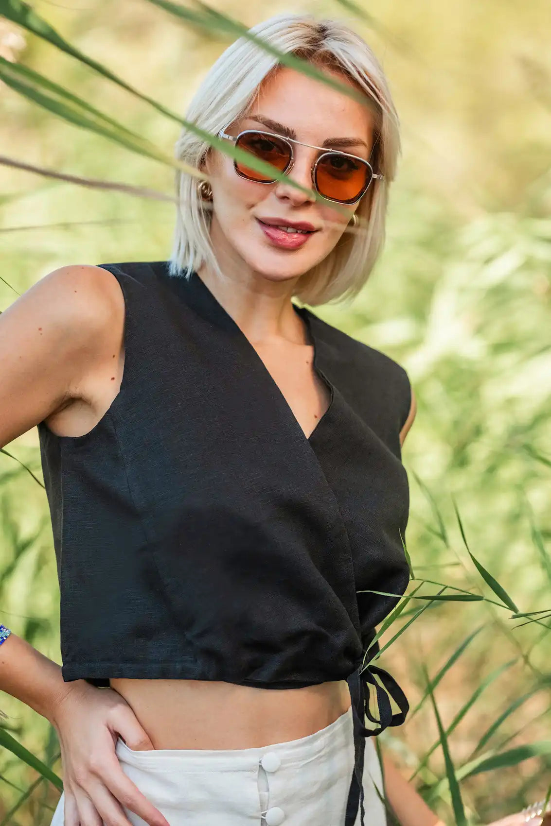 Woman wearing a black sleeveless wrap vest top and sunglasses with a blurred green background
