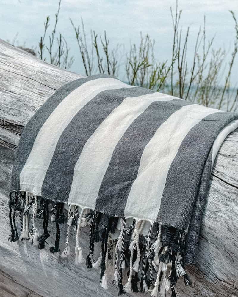 Black and white Striped towel with tassels draped over a log on a beach.