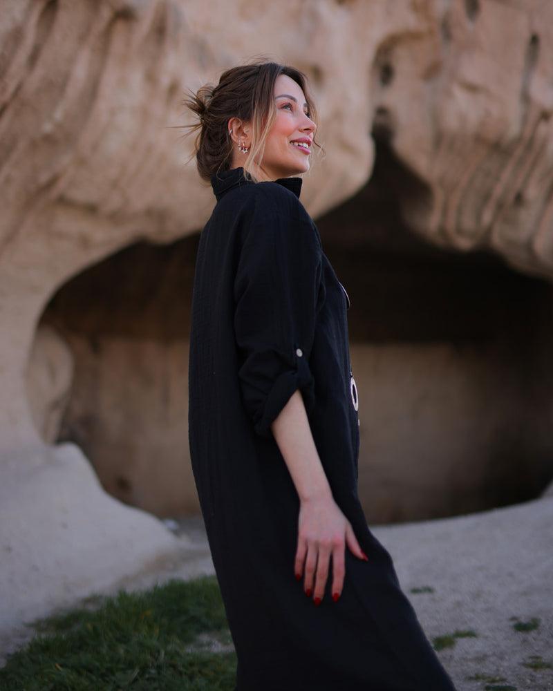 A woman wearing a black dress poses in front of a rock, showcasing her stylish black button-down maxi dress.