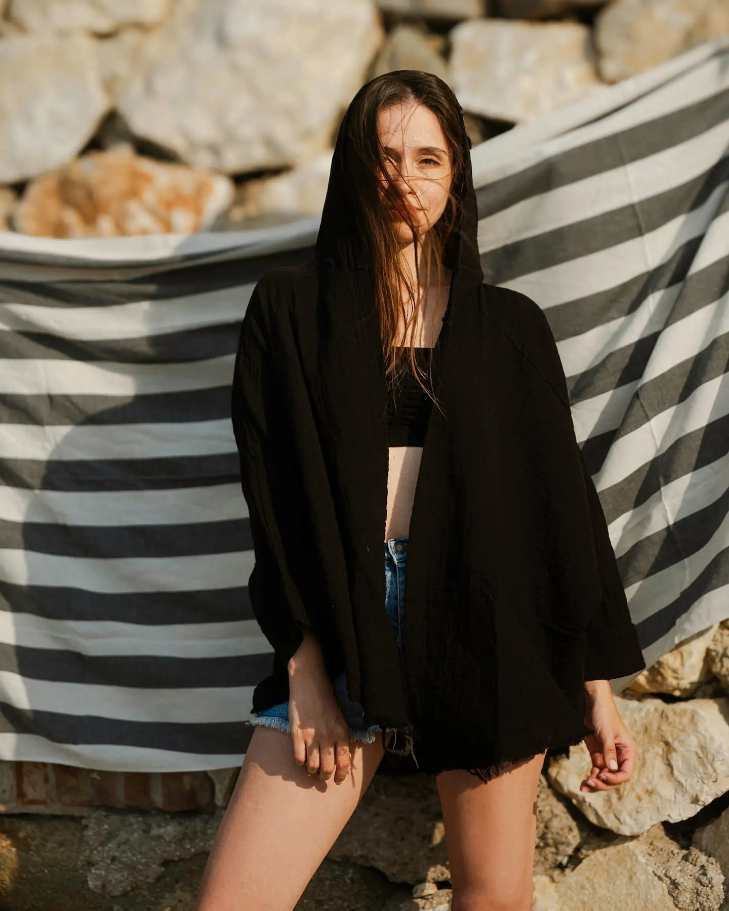 Woman in Black Cardigan with Hoodie standing in front of a striped fabric backdrop with a stone wall background