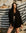 Woman in Black Cardigan with Hoodie standing in front of a striped fabric backdrop with a stone wall background