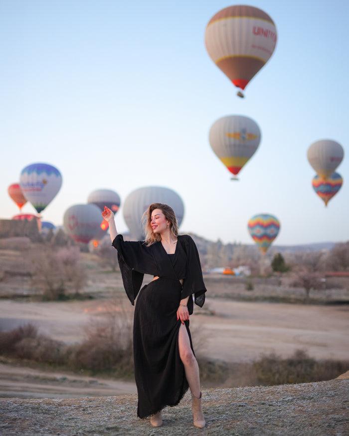 A woman in a black cotton gauze dress stands in front of a colorful hot air balloon against a clear sky.