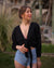 A woman wearing a deep V-neck black wrap top with long sleeves, paired with blue denim shorts and accessorized with a necklace and bracelets.