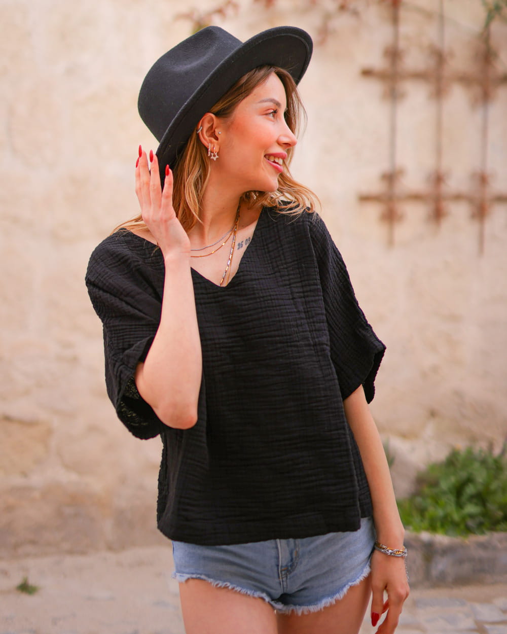 Woman wearing a black hat and black short sleeve cotton top with denim shorts against a stone wall.