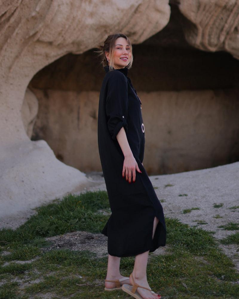 A woman in a black dress and gauze shirt stands confidently in front of a large rock.