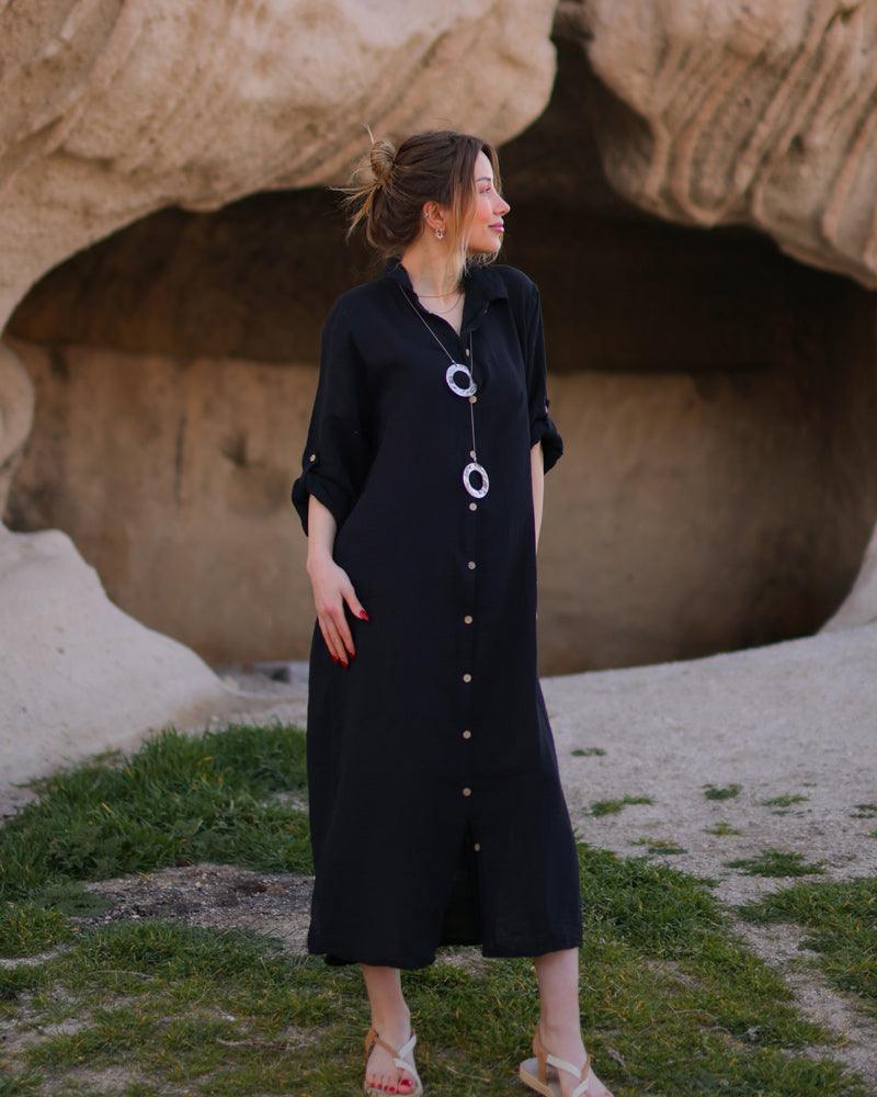 A woman standing outdoors wearing a long black shirt dress with button-up front and long sleeves.