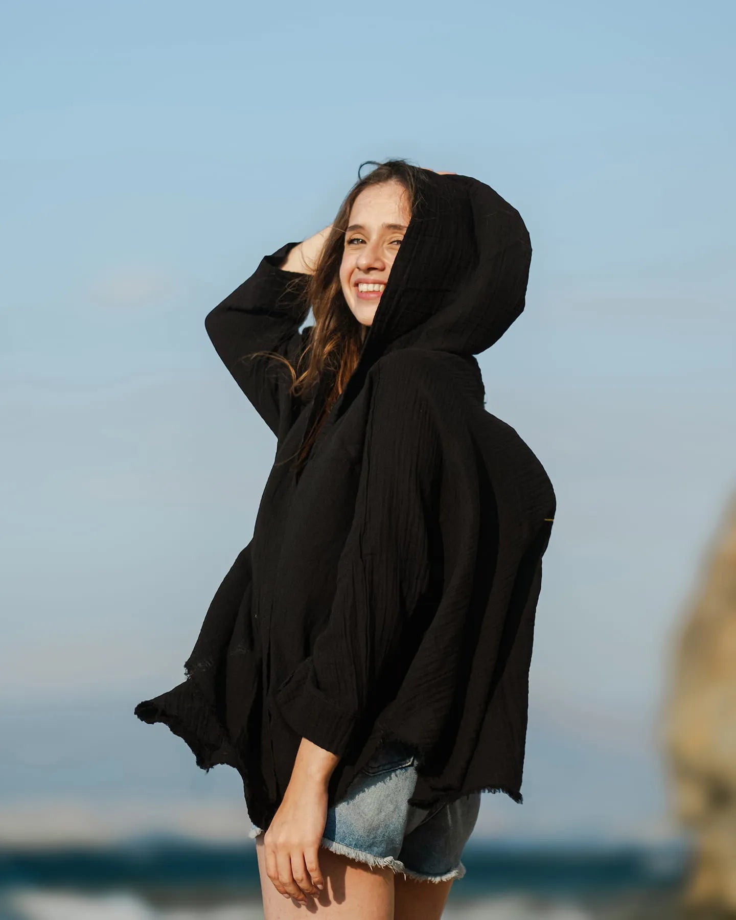 Woman wearing a black hooded poncho on a beach with a clear blue sky.