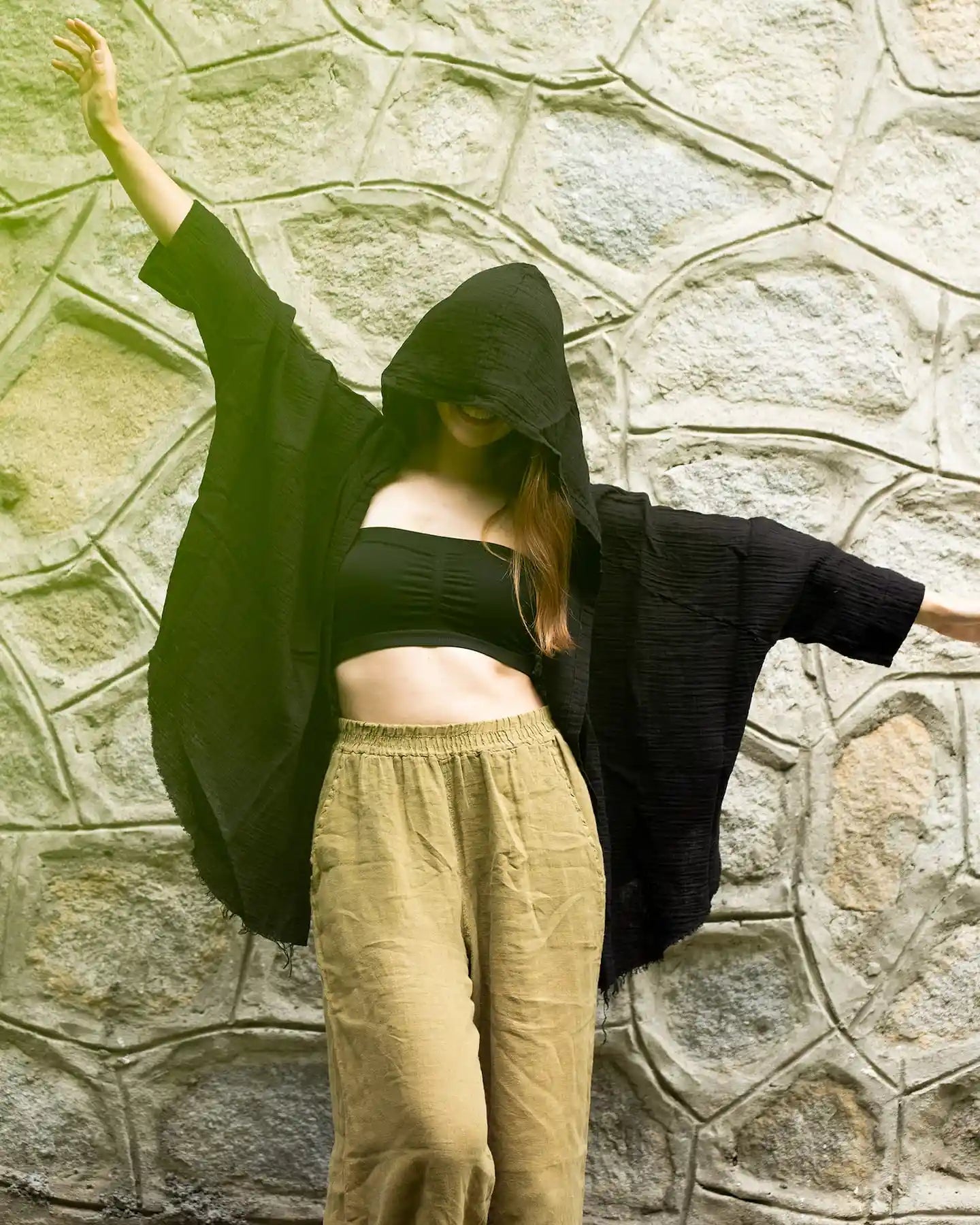 Person wearing a black hooded  double gauze muslin garment against a stone wall
