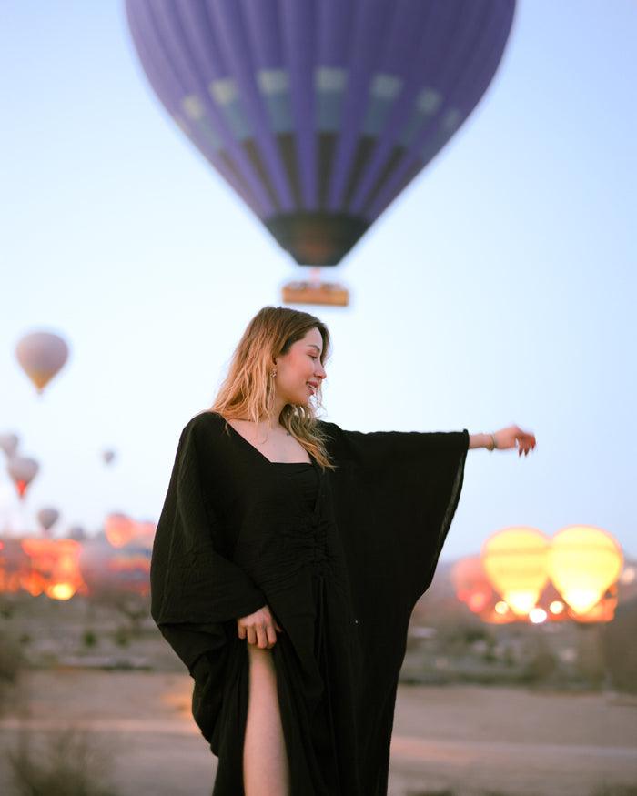  A woman in a soft black kaftan dress stands smiling in front of colorful hot air balloons against a clear blue sky.