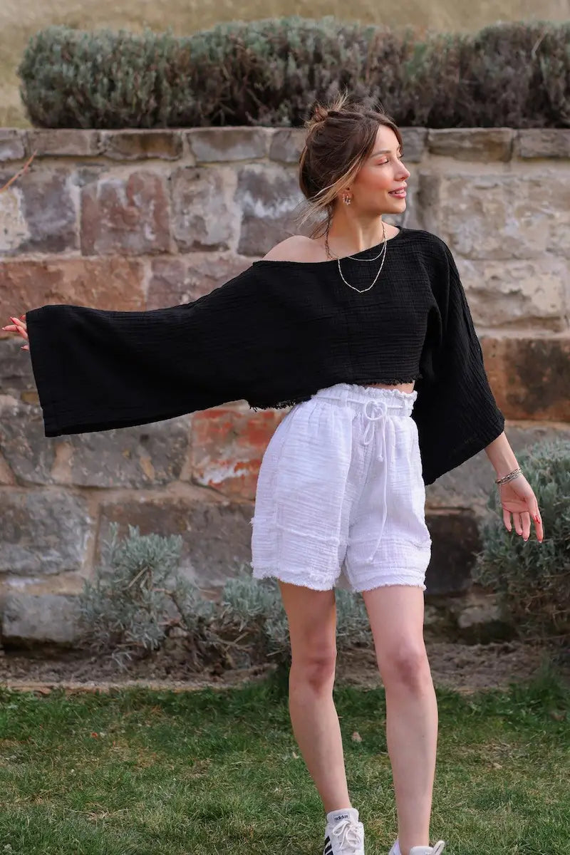 Woman wearing a black off-shoulder cotton top and white shorts standing in front of a stone wall.