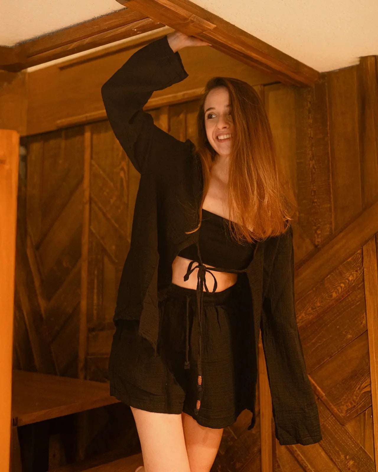 Woman in black open front loungewear top and loungewear shorts standing in a wooden interior setting