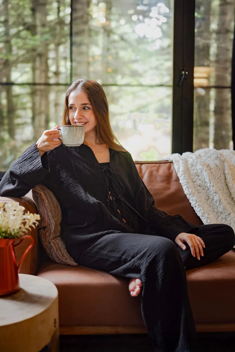Woman sitting in black loungewear set on a couch holding a mug with a forest view outside the window.