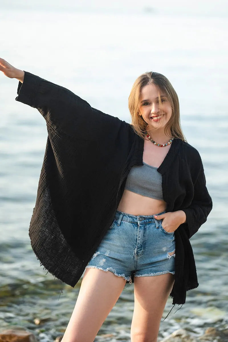 Woman standing by a body of water wearing a black cardigan, gray top, and denim shorts.