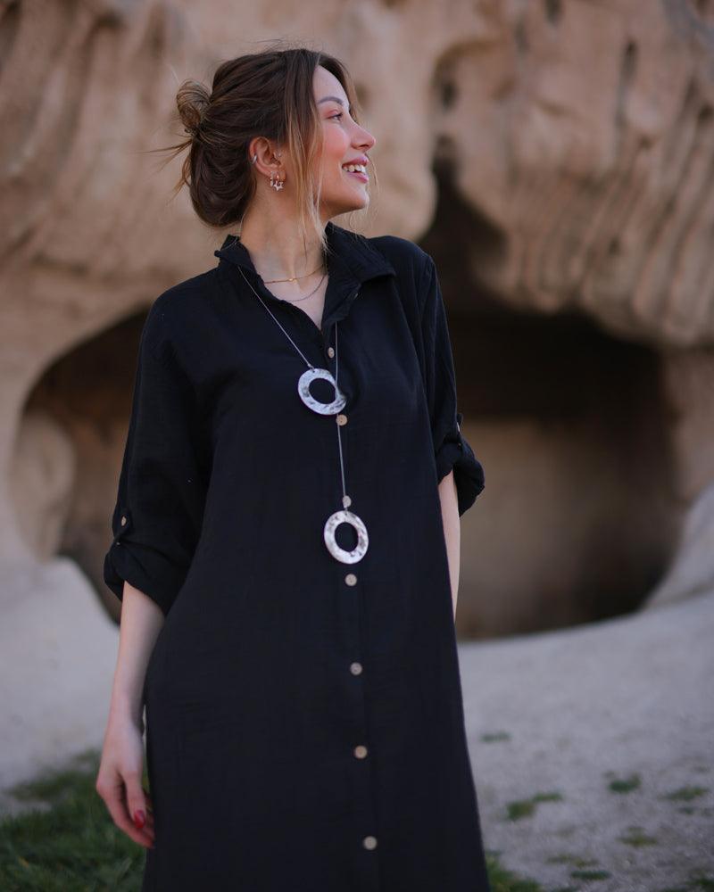 A woman wearing a black shirt dress and a stylish necklace, posing confidently.