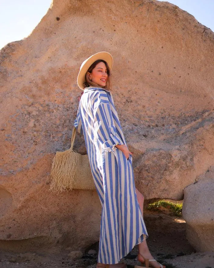 Woman in a blue and white striped shirt dress and straw hat standing against a rocky background