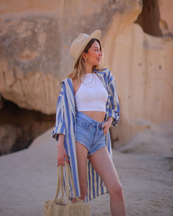 A woman in a blue striped shirt, shorts, and a hat stands in front of a large rock formation.