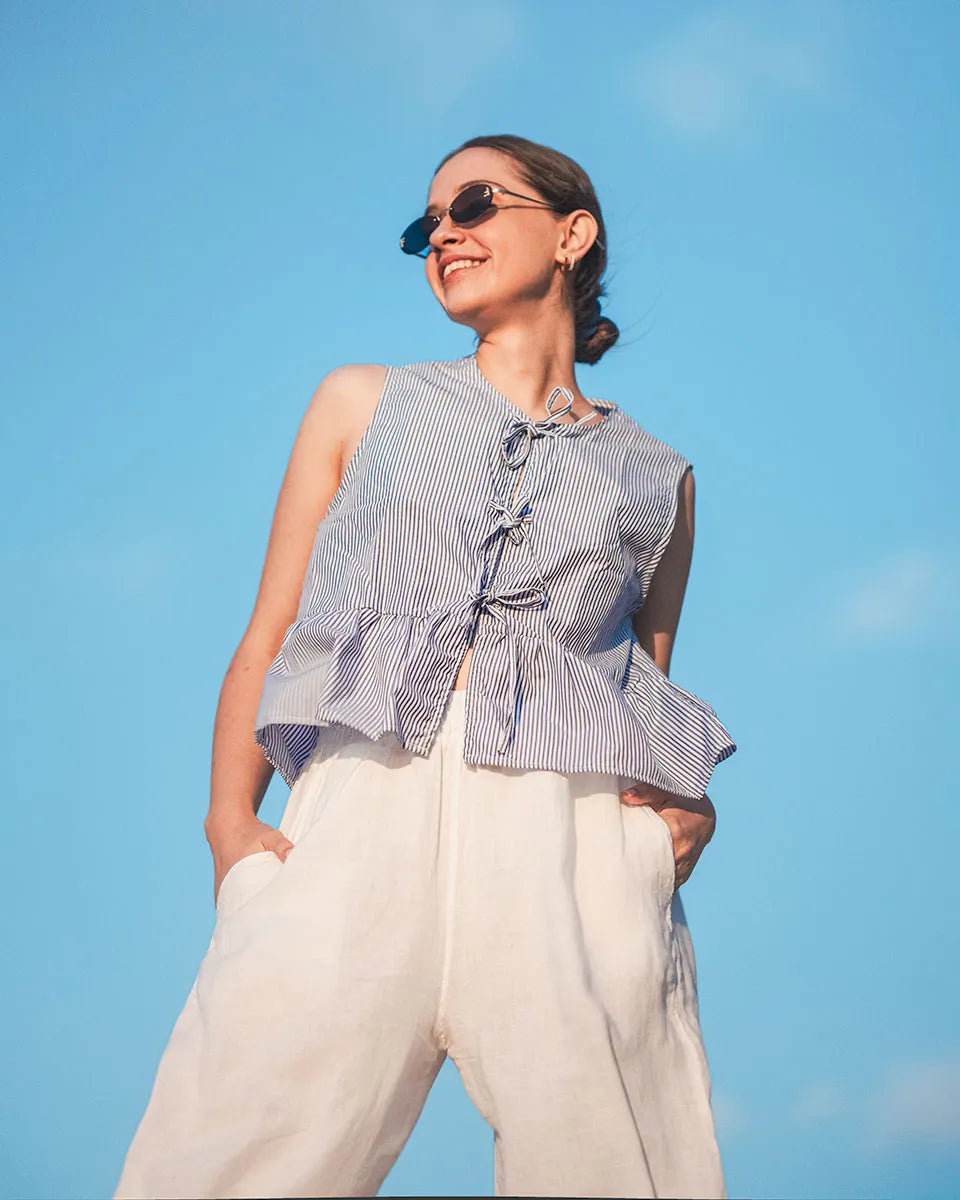Woman wearing a blue striped front tie sleeveless top and white pants against a clear blue sky.