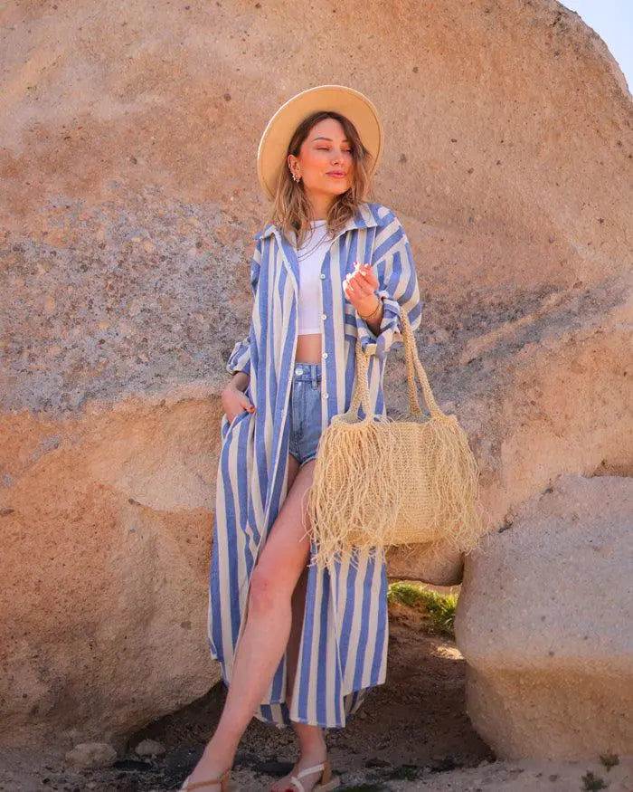 Woman in a striped shirt cover-up and hat holding a woven bag against a rocky background