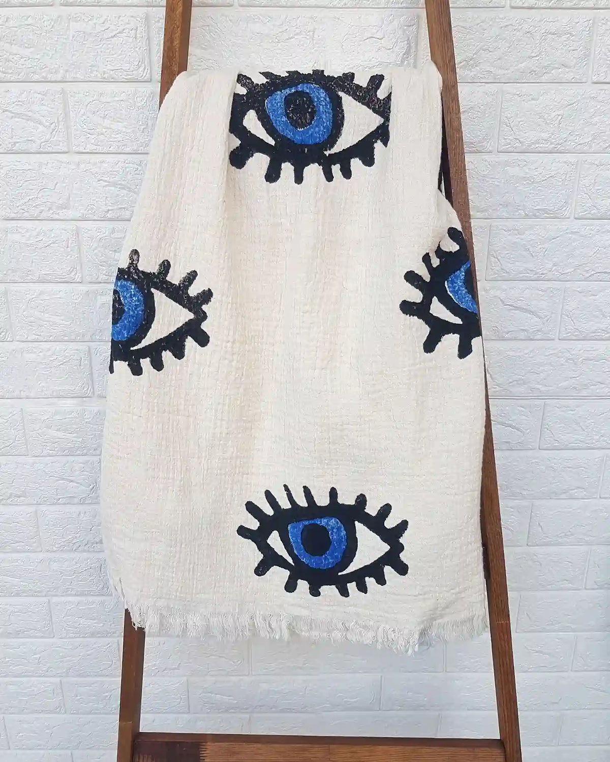 An off-white woven throw blanket with a black and blue eye pattern, draped over a wooden ladder against a white brick wall.