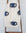 An off-white woven throw blanket with a black and blue eye pattern, draped over a wooden ladder against a white brick wall.