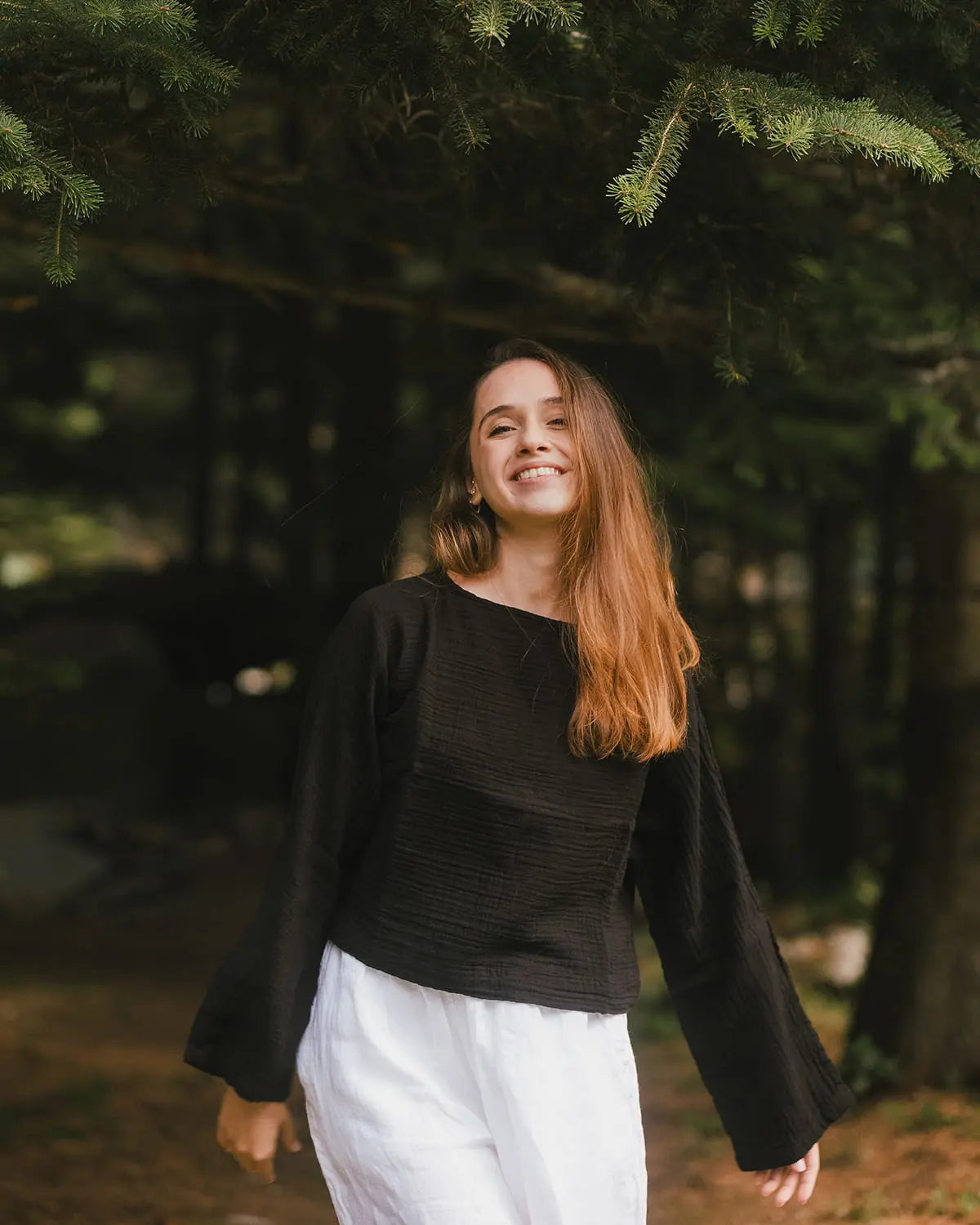 Woman in a black Boat Neck Long Sleeve Top and white pants standing in a forest