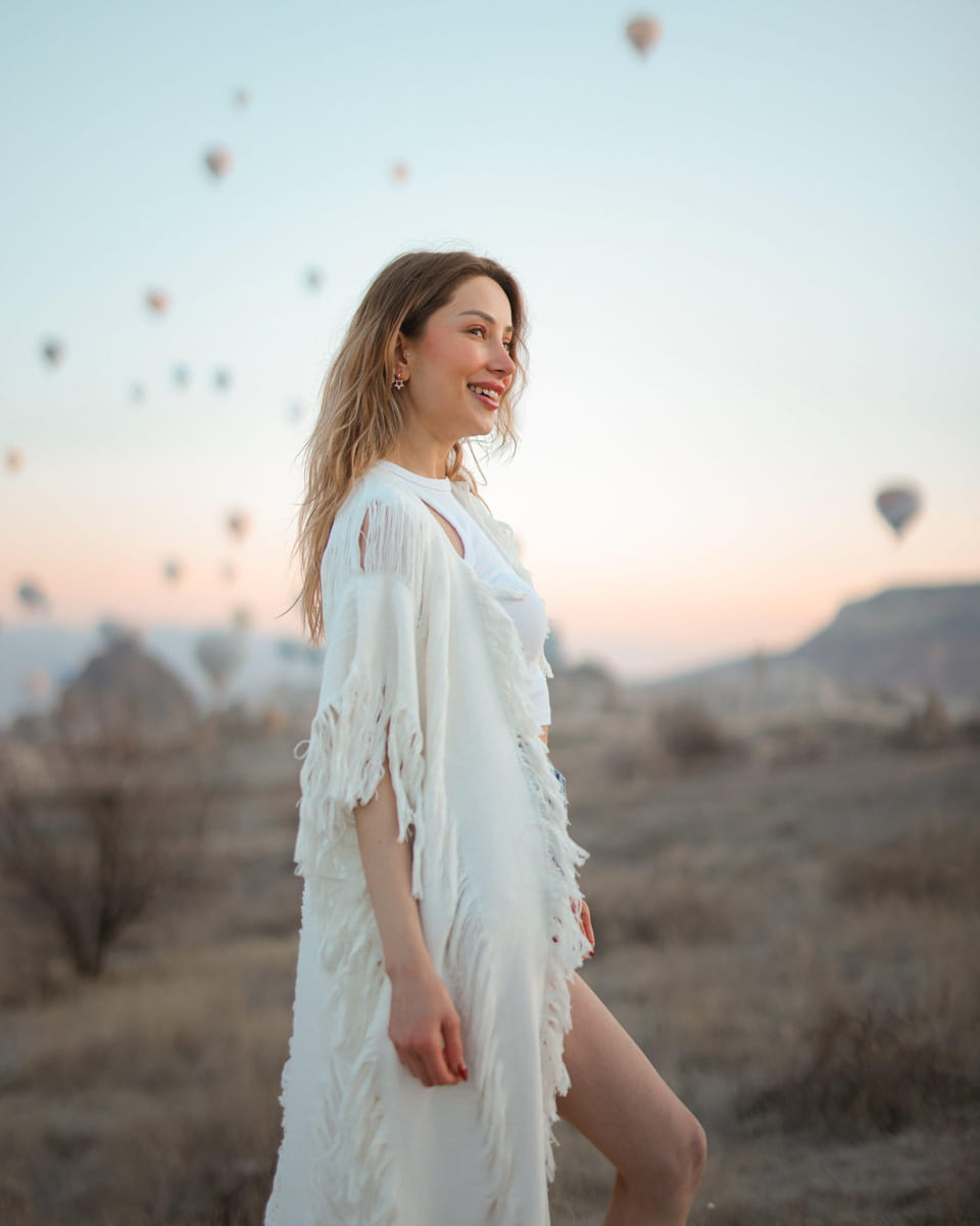 Woman in a bohemian fringed festival outfit dress standing in a desert with hot air balloons in the background