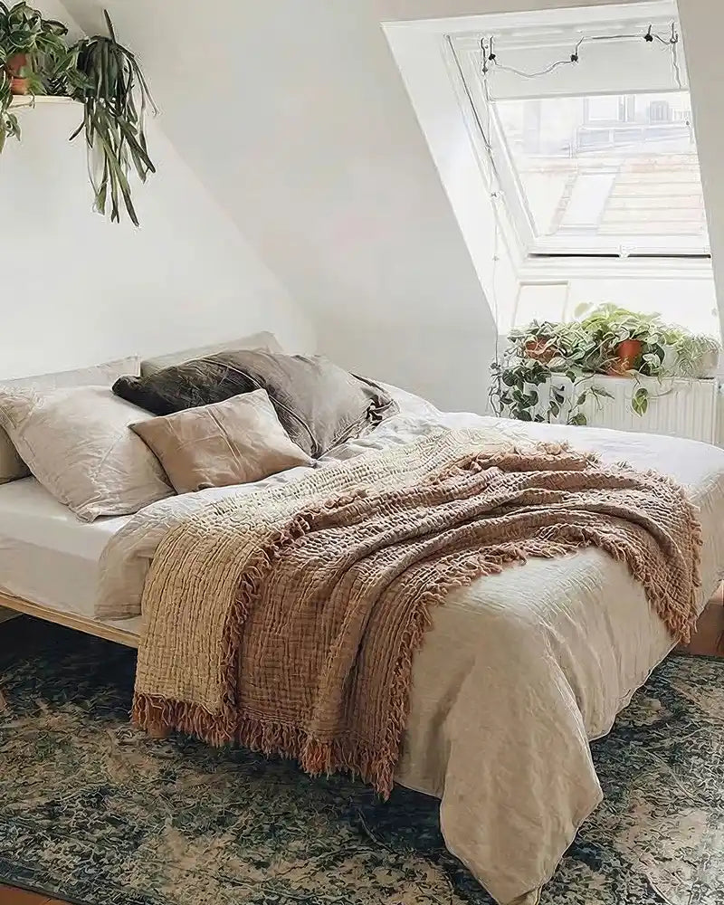 Cozy bedroom with a bed covered in beige and brown blankets, pillows, and a plant on a shelf.