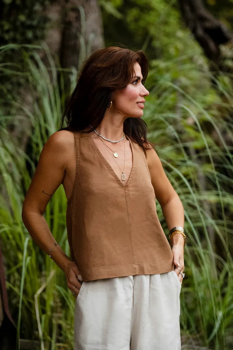 Woman in a brown sleeveless linen top and white pants standing in a natural setting with greenery.