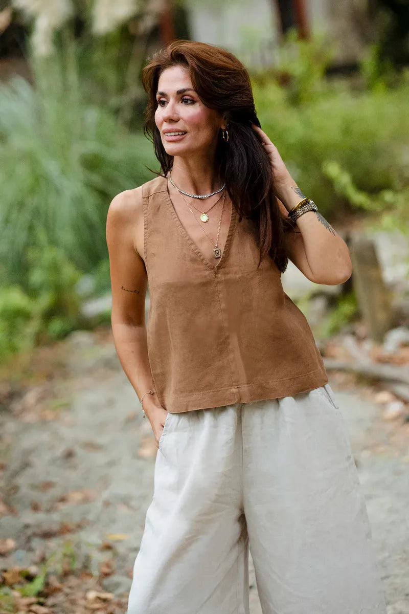 Woman wearing a brown top and light pants standing outdoors with greenery in the background