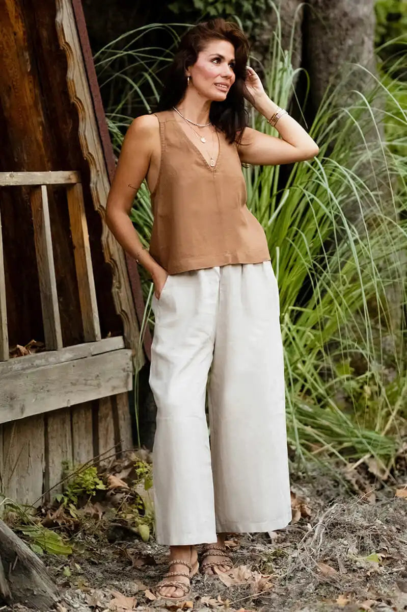 Woman in a brown top and white pants standing outdoors near a wooden structure and plants.
