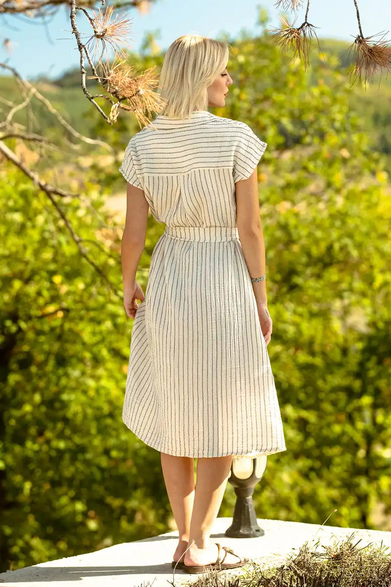 Woman in a striped button down shirt dress standing outdoors with greenery in the background