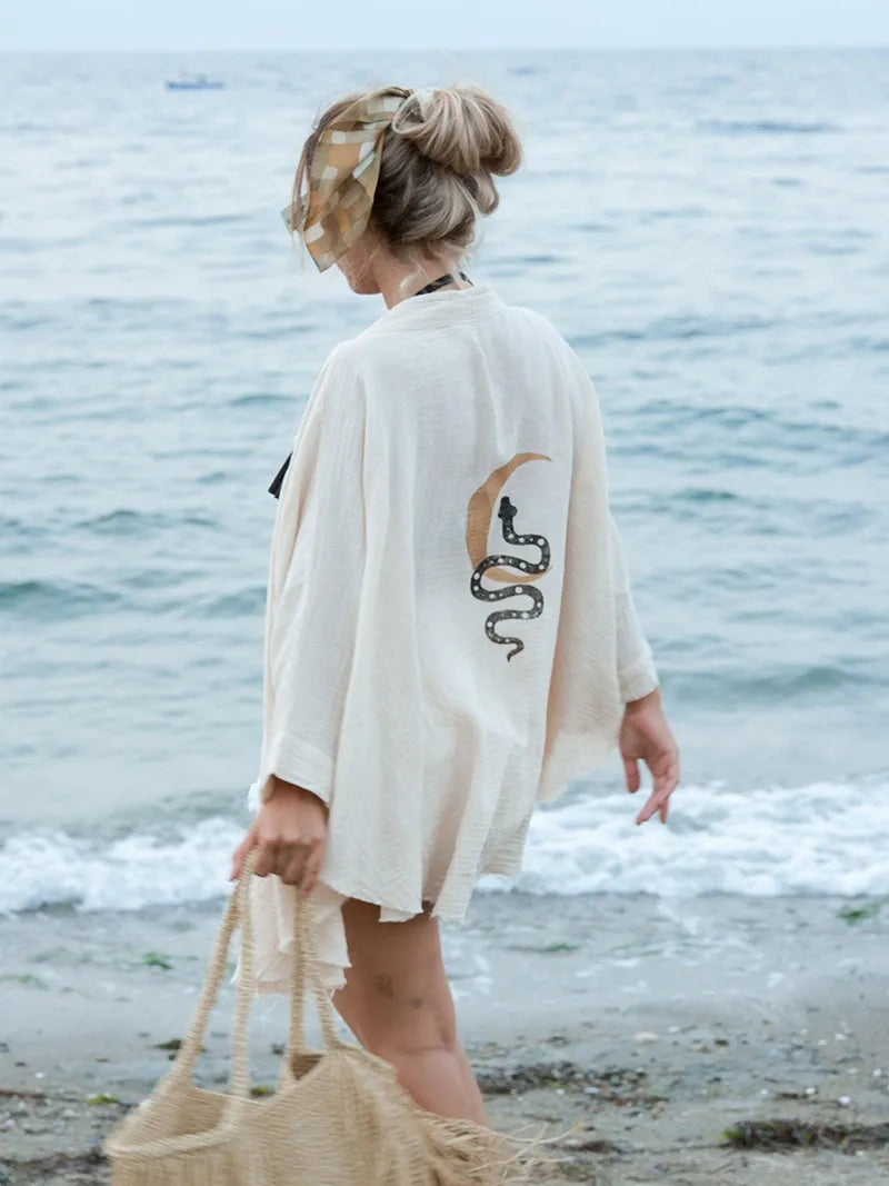Woman standing on a beach wearing a beige kimono with a a crescent moon and snake design, holding a woven bag.