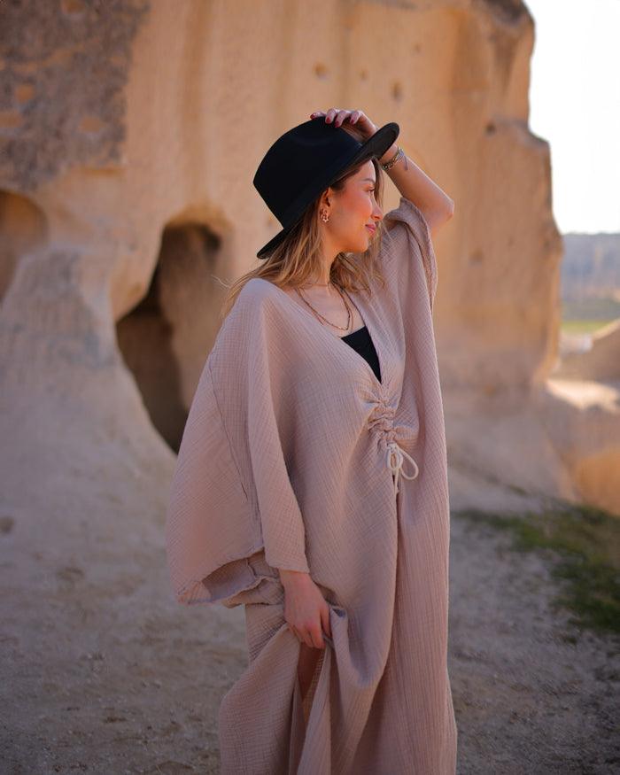 A woman in a flowing cotton gauze dress and hat stands against a backdrop of rugged rock formations.