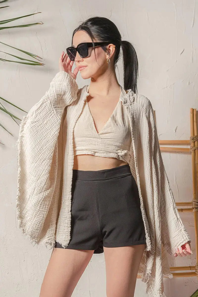 Woman wearing sunglasses and a cream muslin kimono top over a crop top and shorts against a light background