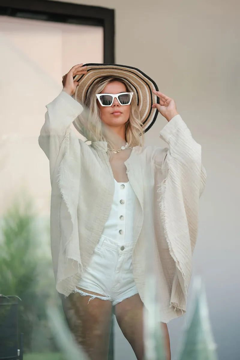 Woman wearing a cream long sleeve muslin robe with a wide-brimmed hat and sunglasses, standing outdoors.
