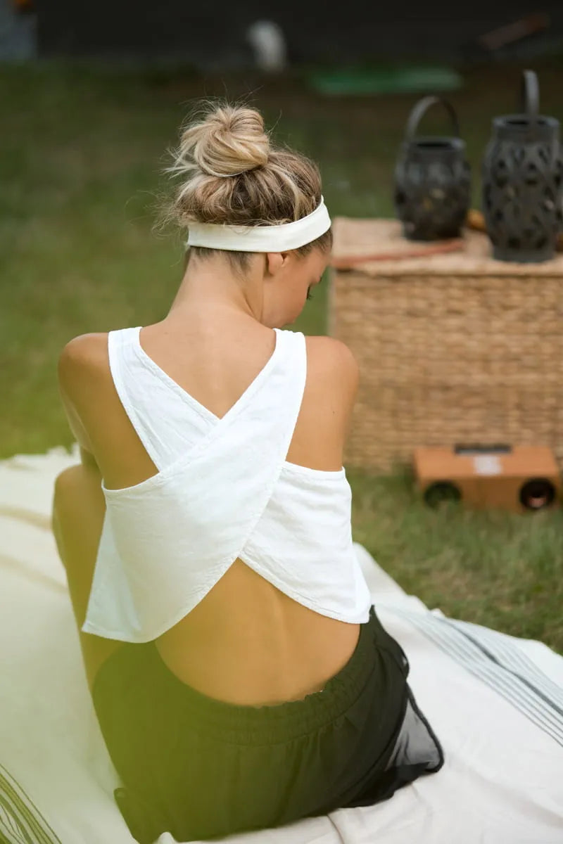 Woman sitting on a blanket outdoors wearing a white top with a criss-cross back design.