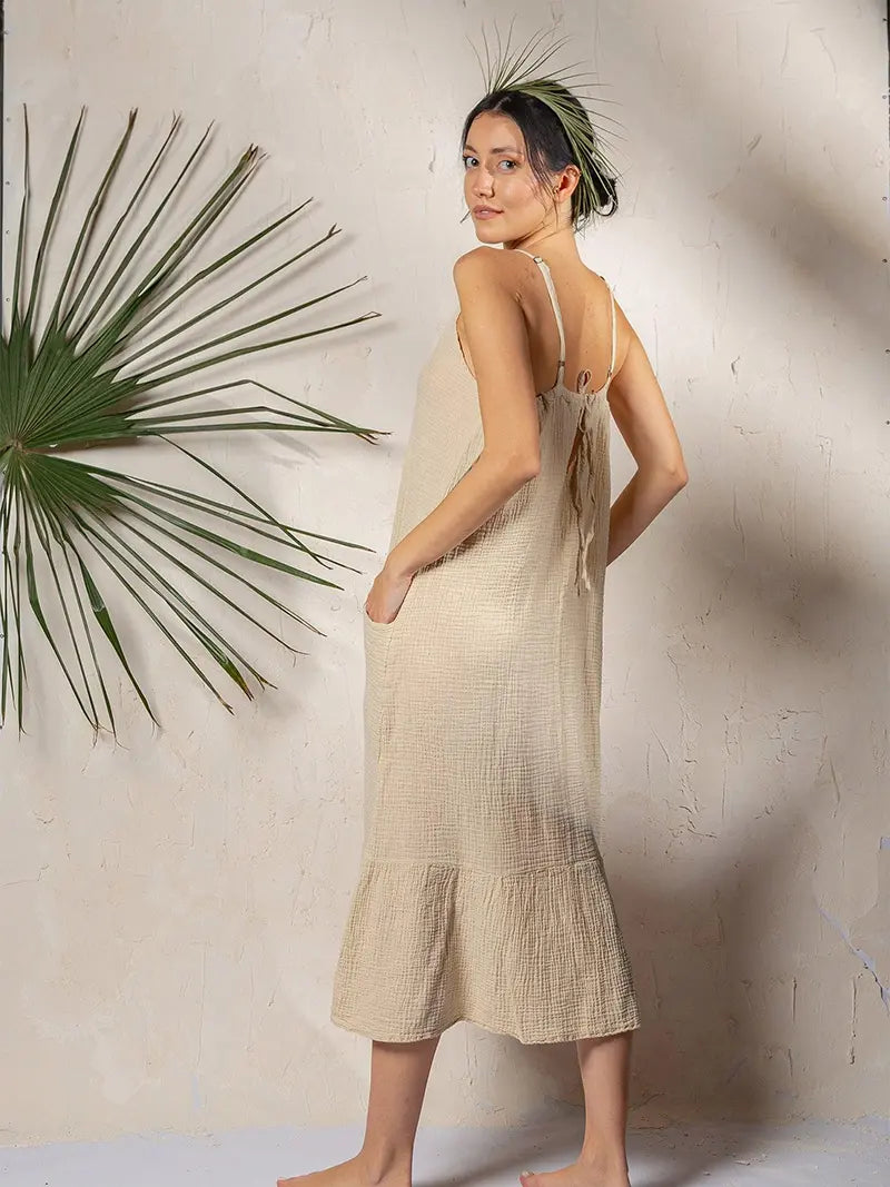 Woman wearing a beige soft cotton muslin dress standing against a textured wall with a plant.