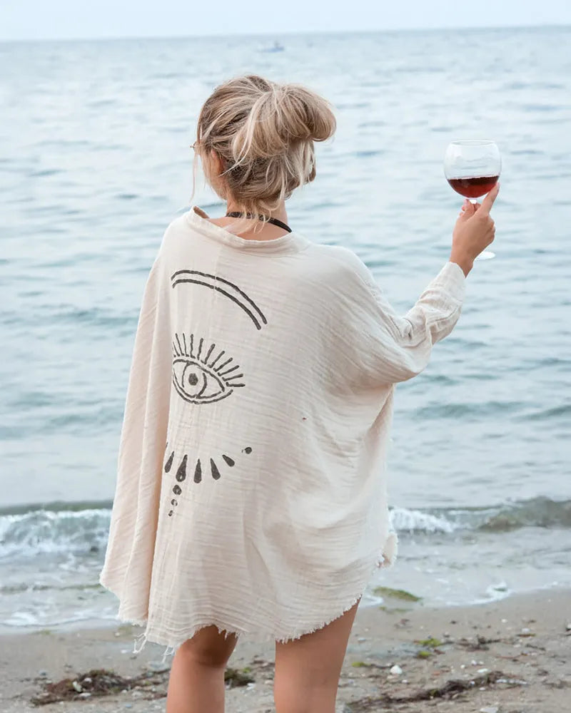 Woman on a beach holding a glass of red wine, wearing a cream cover-up with an eye design.