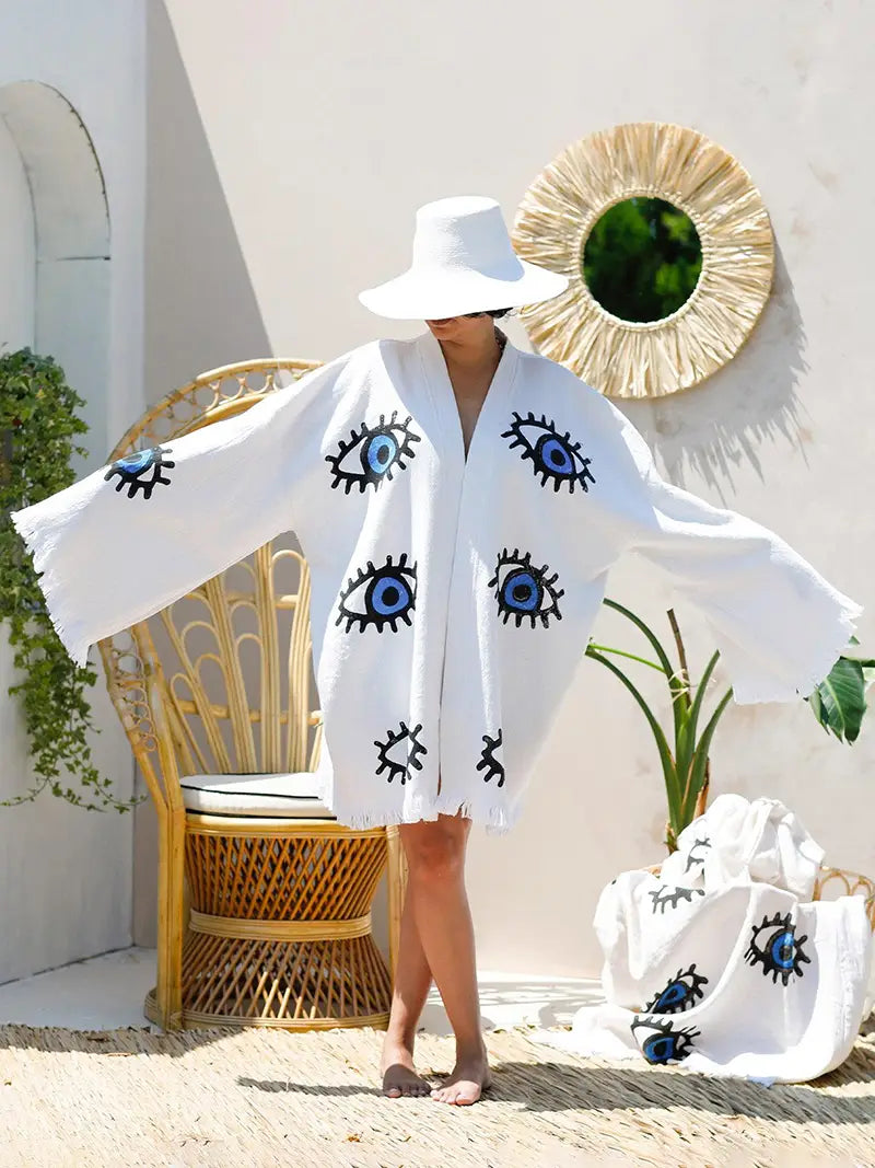 Person wearing a white kimono cover-up with evil eye patterns in a sunlit outdoor setting.