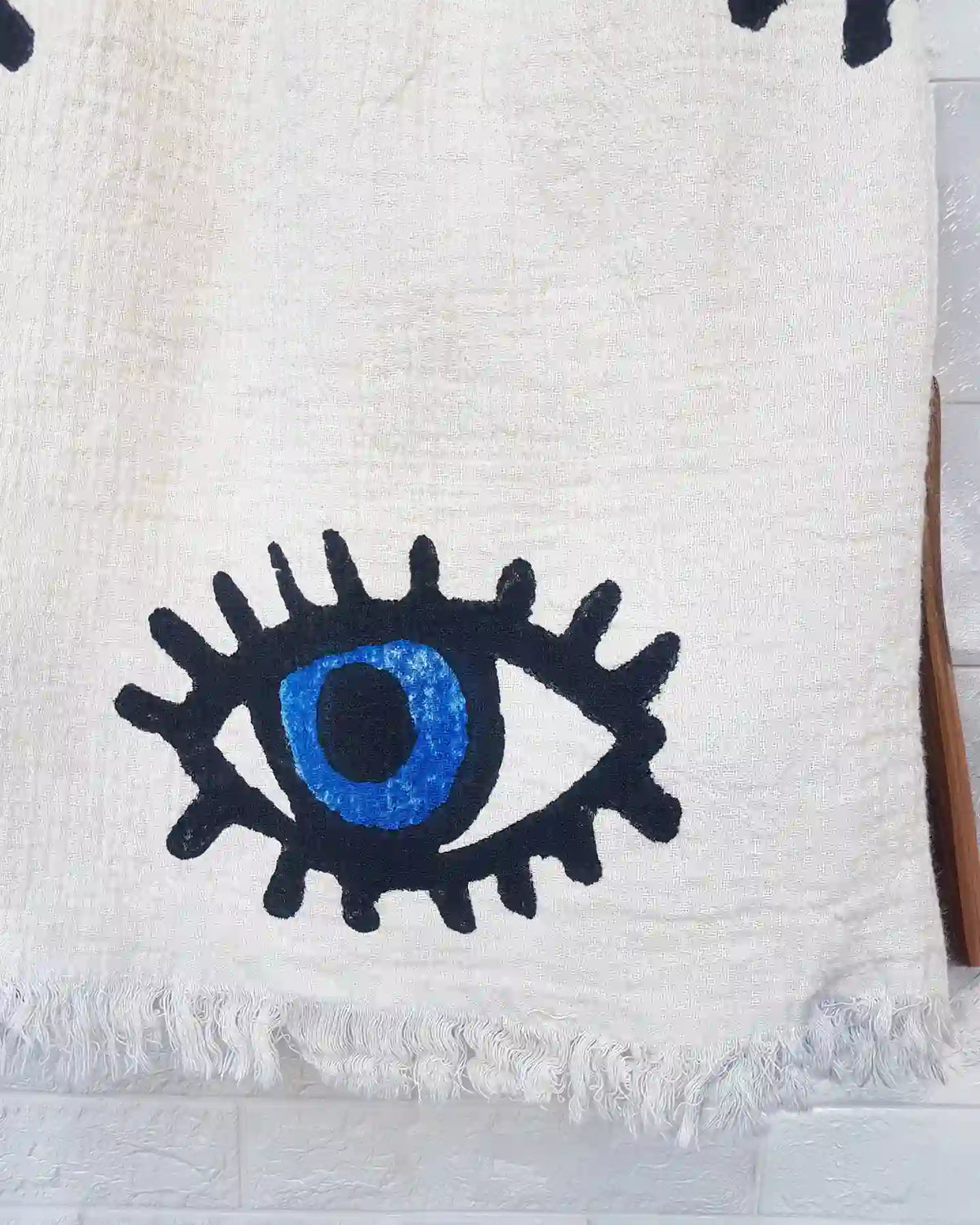 A close-up of a white woven textile with a large stylized eye motif in black and bright blue, plus fringed edges, shown against a light wall.
