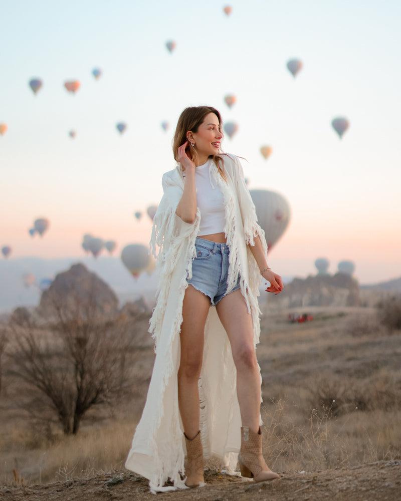 A person wearing a white frayed kimono with long sleeves, styled with a white top and blue denim shorts, standing in a dry landscape with hot air balloons in the background.