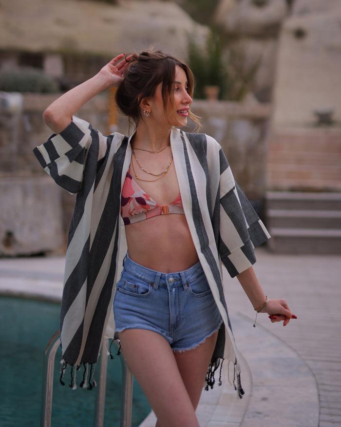 A woman is positioned in front of a pool, wearing a fringed striped Turkish robe