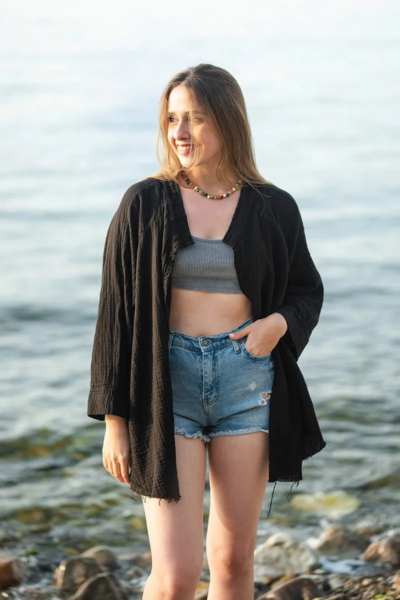 Woman standing by the water wearing a black cardigan, and denim shorts.